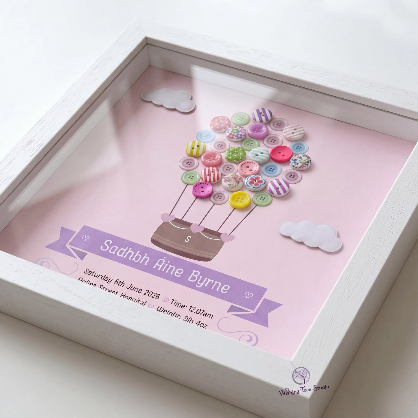 A square white shadow box frame containing a personalised pink print with a hot air balloon made from colourful buttons, clouds, and details for a new baby below.