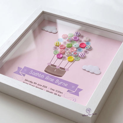 A square white shadow box frame containing a personalised pink print with a hot air balloon made from colourful buttons, clouds, and details for a new baby below.